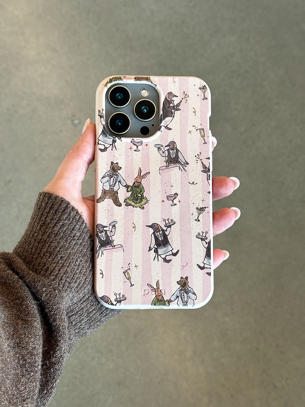 A hand holding the Pela Cocktail Cabana Phone Case, featuring a whimsical design of animals dressed for a party—penguins, bears, and rabbits—enjoying cocktails on a pink and cream striped background.