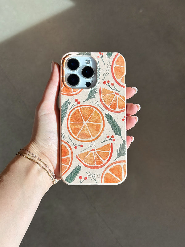 Hand holding the Pela Citrus Cheer Phone Case featuring a festive design of orange slices, green pine branches, and red berries on a cream background.